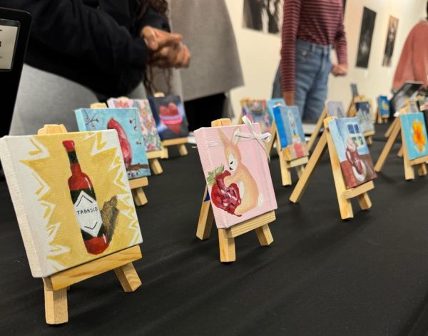 Mini canvases showcasing student artwork, including a Tabasco bottle painting with vibrant yellow zigzags and a furry animal with its strawberry, stand displayed on wooden easels at Jefferson's NAHS holiday art sale. “I think the [artists] at our school are extremely talented. This time I could walk through and really admire the skill these [students] have,” Senior Alex Do said.